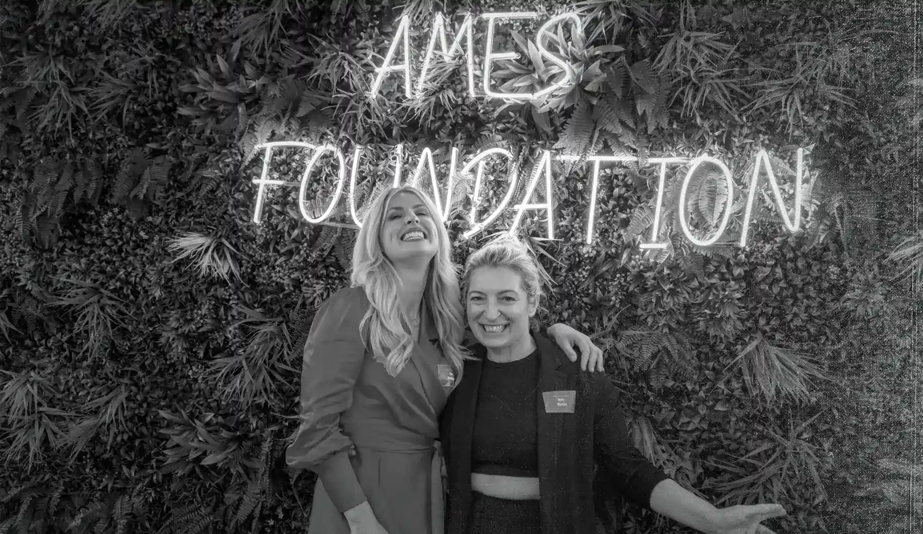 Black and white picture of smiling Lea C. Henzgen hugging a friend infront of plant wall and neon AMES-Foundation sign